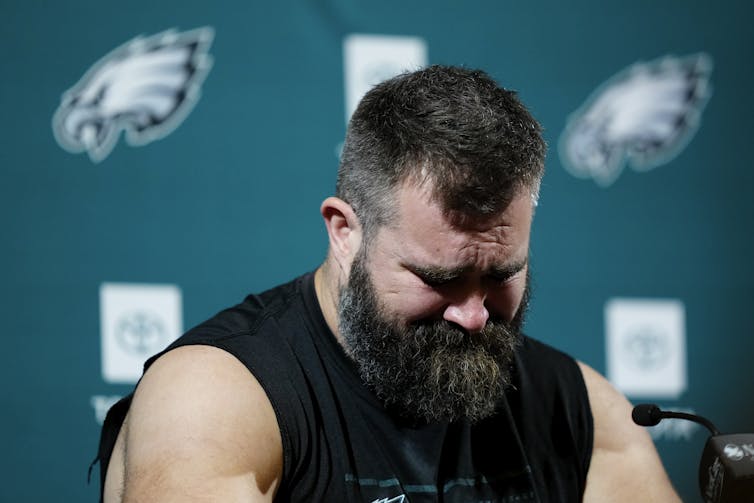 A dark-haired, bearded man in a black T-shirt with cut off sleeves looks sad.