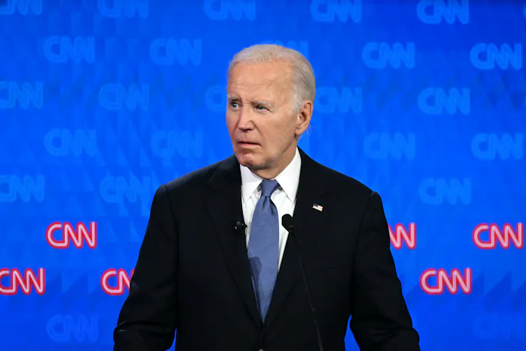 A gray-haired man in a dark suit, standing on a stage, looks at something to his right.