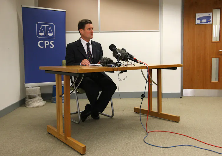 A young Keir Starmer sitting behind a table giving a press conference in front of a CPS banner.