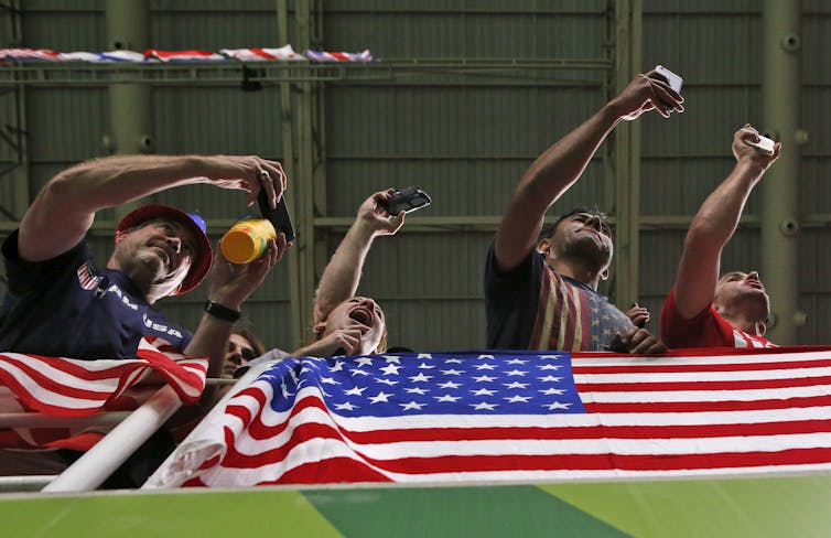 Four people with their arms extended, cheering, seen from below with an American flag in the foreground.