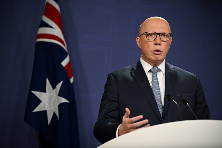 A man with glasses and a suit stands in front of a lectern, behind him an Australian flag