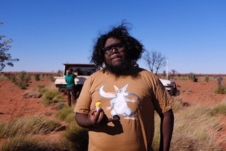 A person with black curly hair in a t-shirt with a bilby cartoon stands in the red outback holding a small plastic vial.