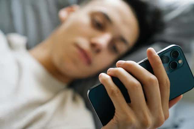A young man lies on a bed, looking at his phone. 