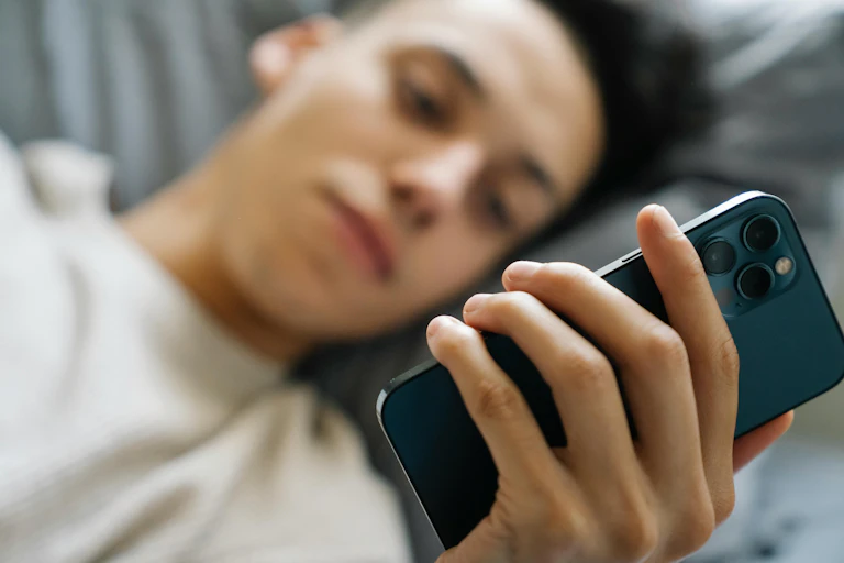 A young man lies on a bed, looking at his phone.