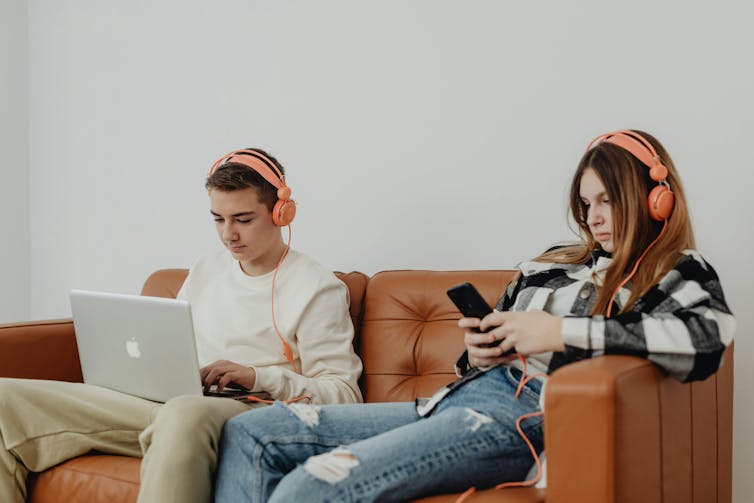 A teenage boy and girl sit side. by side on a couch. Both wear headphones. The boy types on a laptop, then girl looks at a phone.