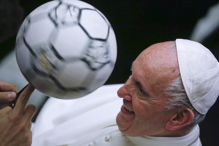 A man wearing a white gown and white cap looks intently at a soccer ball.