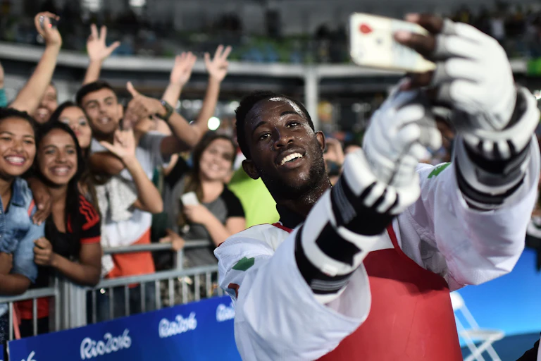 A grinning athlete in protective gloves and chest guard holds up a phone to take photos with a group of grinning people