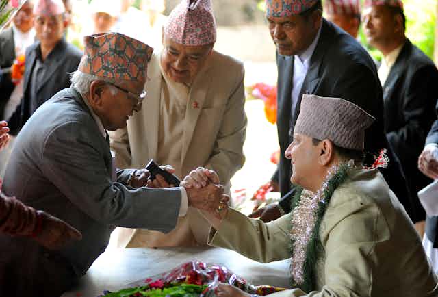 A seated man with a flower garland around his neck clasps the hand of another, while several men look on.