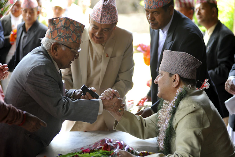 A seated man with a flower garland around his neck clasps the hand of another, while several men look on.