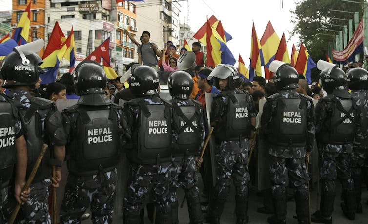 People in helmets and jackets that say 'Nepal police' block off a group on top of a van holding flags with red, yellow and blue colors.