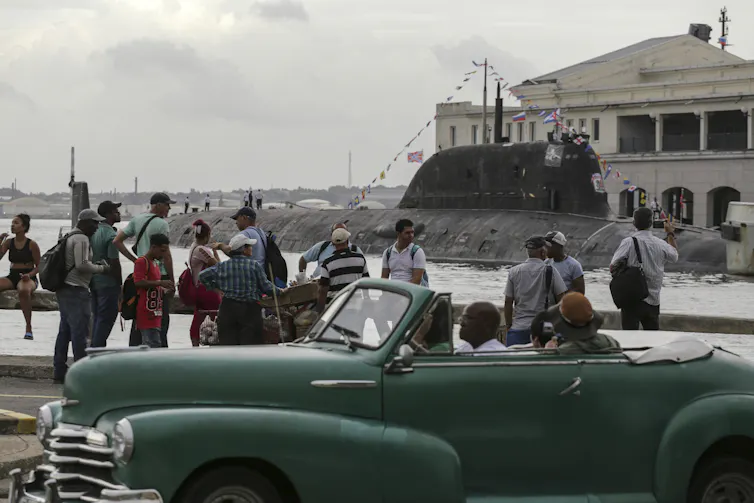 people along a waterfront watch as a submarine sails by.