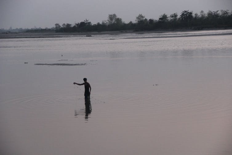 A man stands in a low river with a fishing net in his hand.