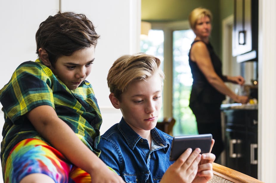 An out-of-focus woman in the background looks on as two boys stare at a screen.