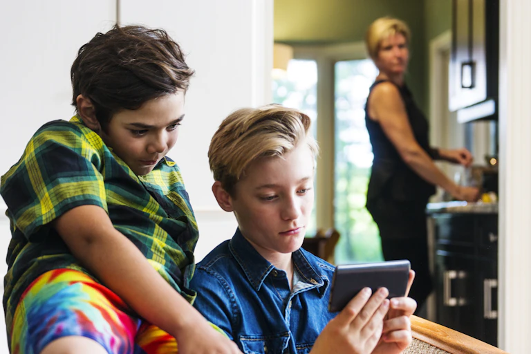 An out-of-focus woman in the background looks on as two boys stare at a screen.