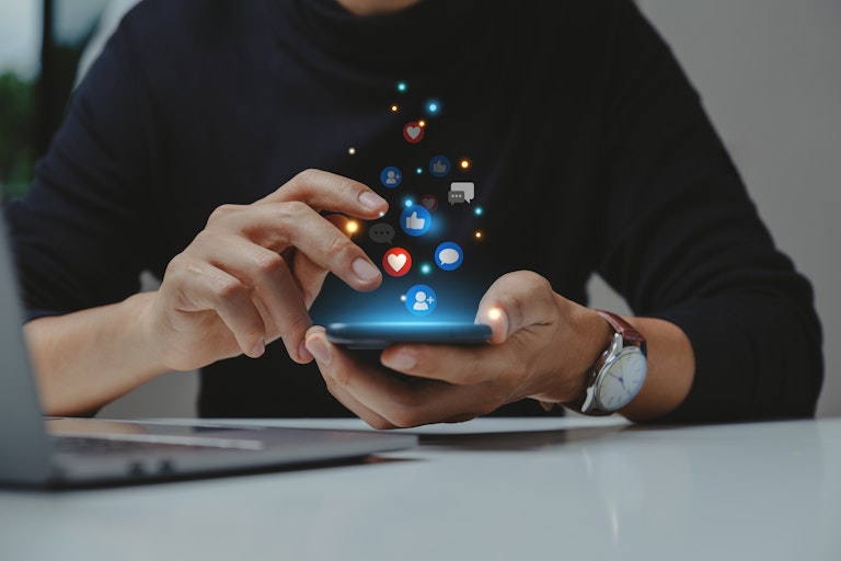 A person's hands close up on a smartphone with reaction emojis floating above it.