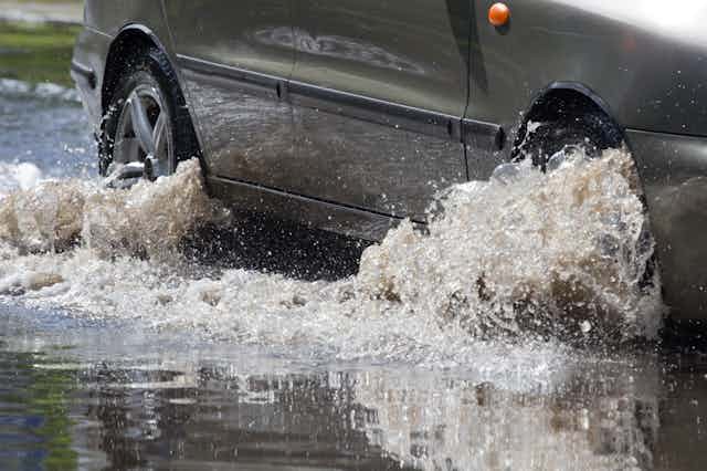 Closeup of a car driving on a flooded road, with shallow water churning around the wheel hubs 