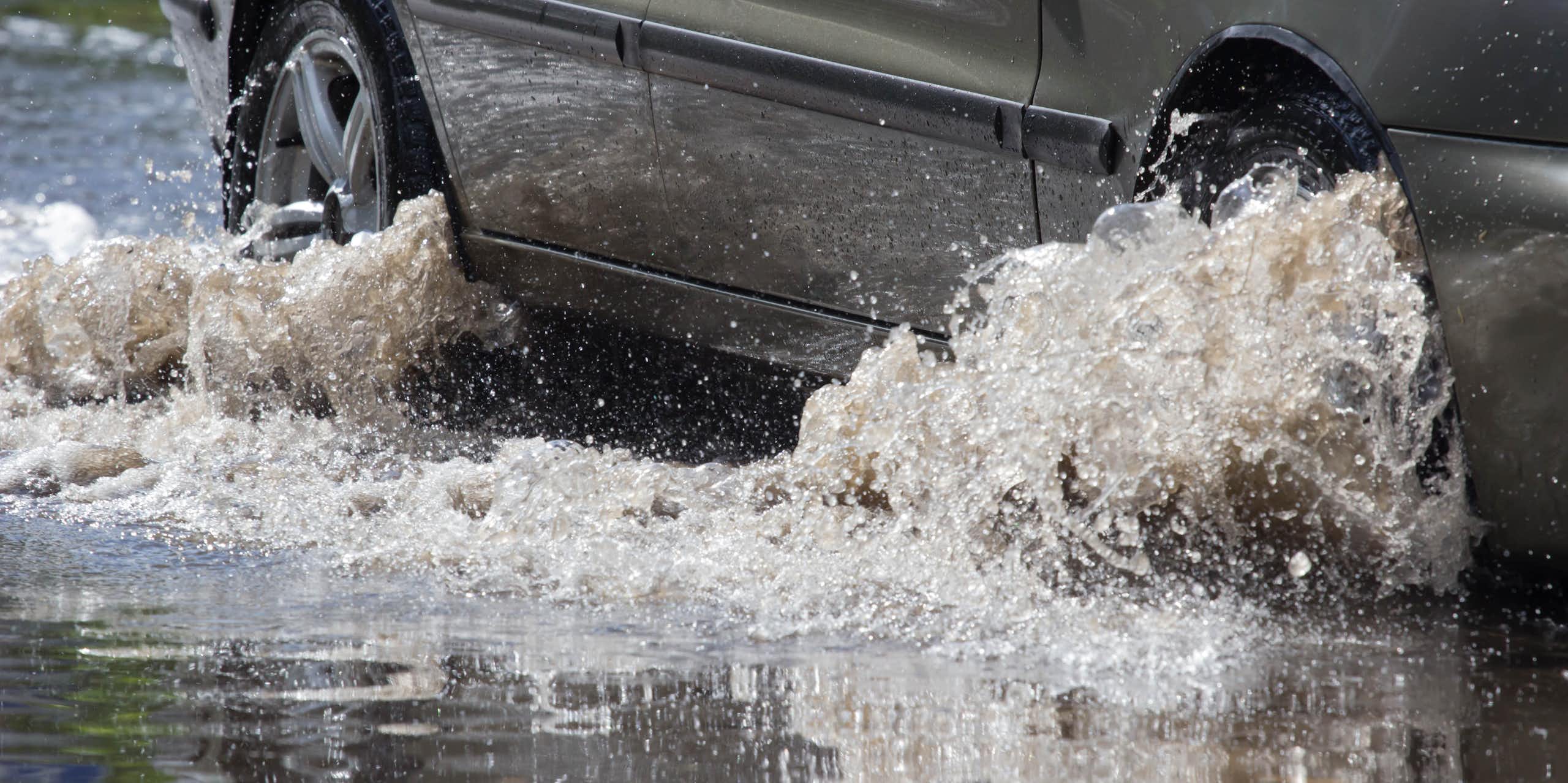 Closeup of a car driving on a flooded road, with shallow water churning around the wheel hubs 