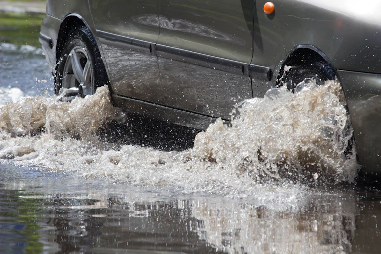 Closeup of a car driving on a flooded road, with shallow water churning around the wheel hubs