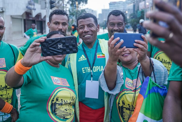 A grinning man in athletic clothing poses with several people using their phones to take selfies