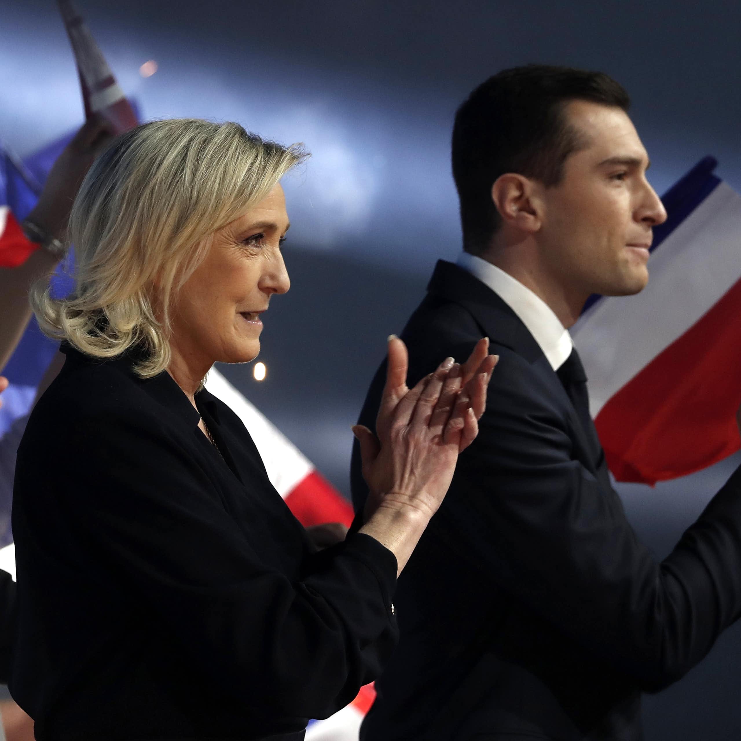 Marine Le Pen and Jordan Bardella stood in front of France flags applauding a crowd.