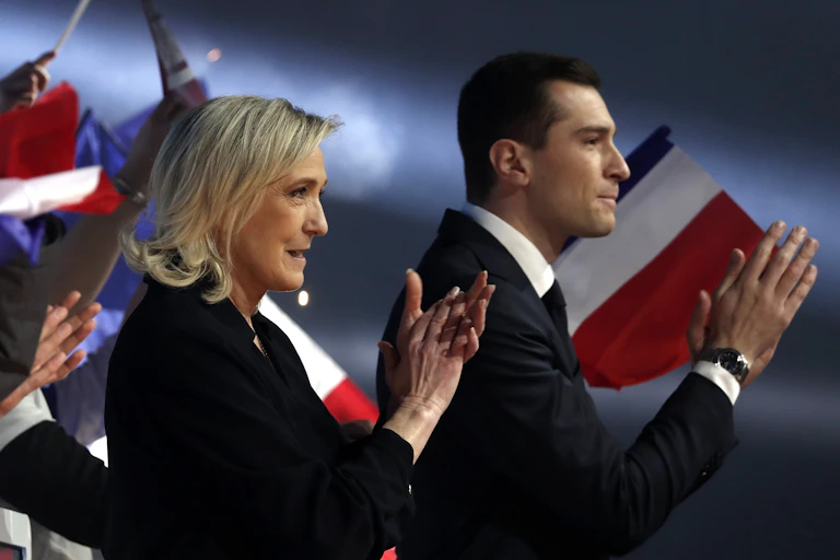 Marine Le Pen and Jordan Bardella stood in front of France flags applauding a crowd.