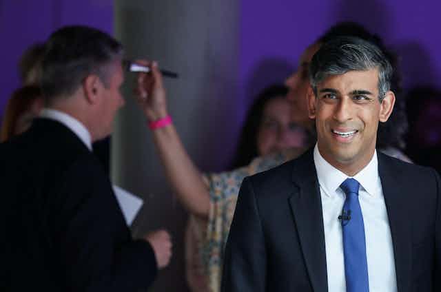 Rishi Sunak smiles before his election debate with Keir Starmer, who receives makeup in the background.