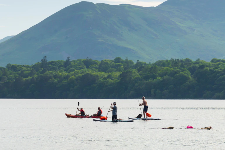 Lake district scene with two paddleboards, two kayakers and swimmers in foreground on water