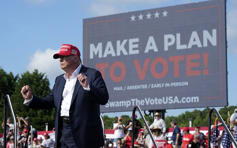 A man in a red ball cap with his hands raised and clenched in fists stands in front of a sign that reads Make a Plan to Vote.