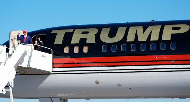 A man in a blue suit wearing a red ball cap raises his fist while disembarking from a plane that as a large Trump on its fuselage.