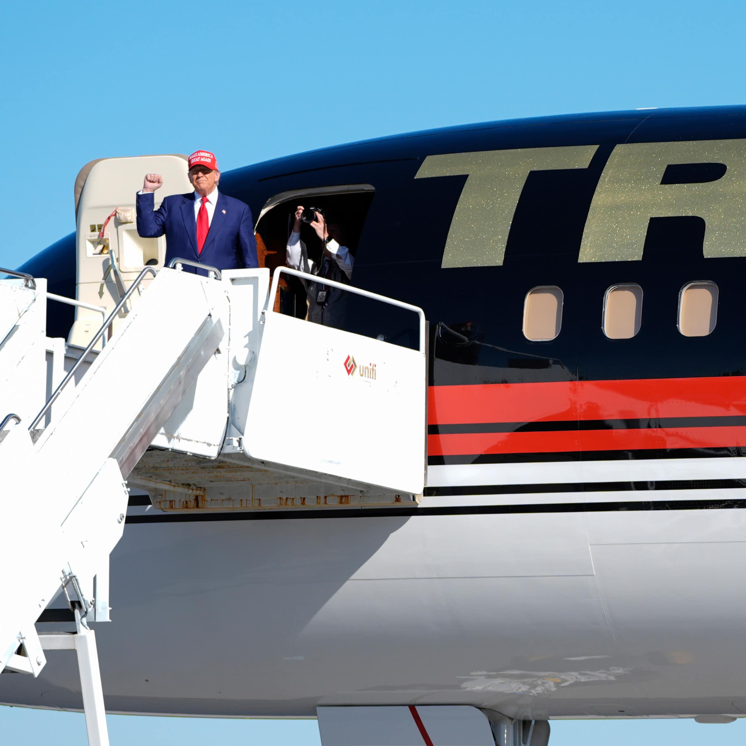 A man in a blue suit wearing a red ball cap raises his fist while disembarking from a plane that as a large Trump on its fuselage.