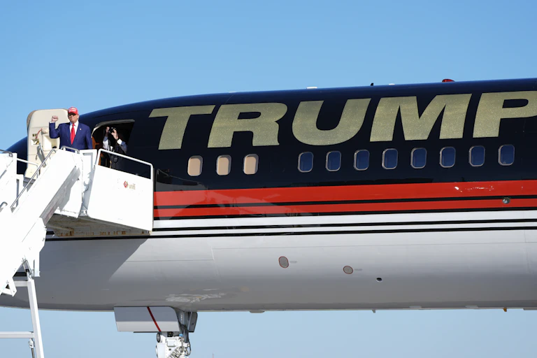 A man in a blue suit wearing a red ball cap raises his fist while disembarking from a plane that as a large Trump on its fuselage.
