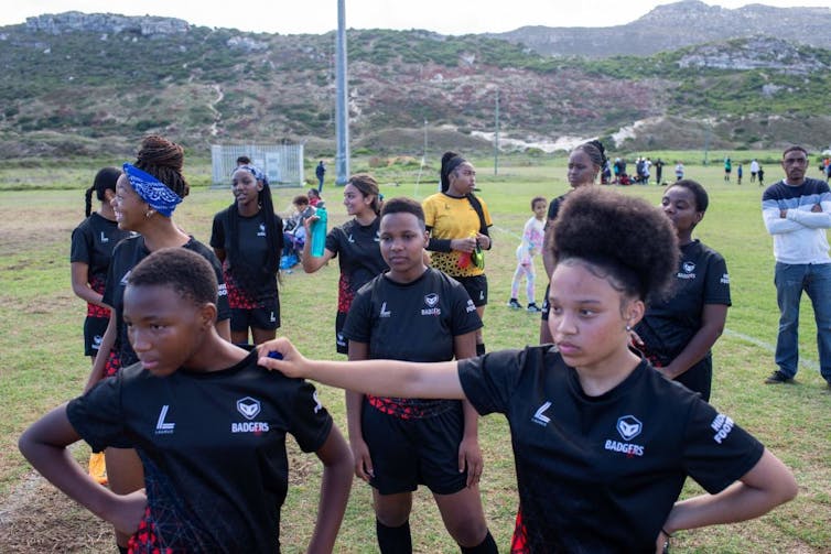 A group of girls in black sports uniforms are gathered on a sports field.  In front, a girl with an Afro rests her hand on another girl's shoulders.  There are mountains in the background.