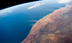 Satellite image showing the blue of the south Atlantic on the left and then the Namib Sand Sea, with its distinctive orange-red sand, and then inland to the Great Escarpment mountains. Plumes of dust are seen being blown from the dry river valleys.