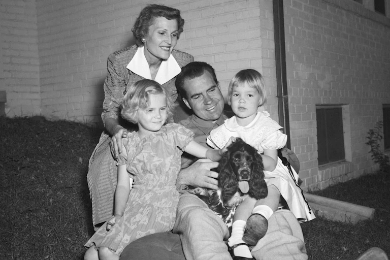 A man sits with two small children and a dog on his lap as a woman stands behind them and smiles.