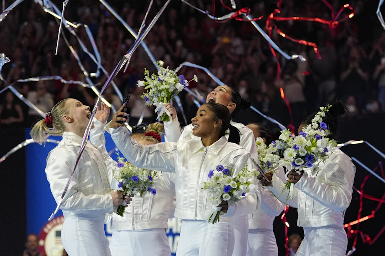 Young women dressed in white slacks and jackets laugh as they look up while holding flowers.