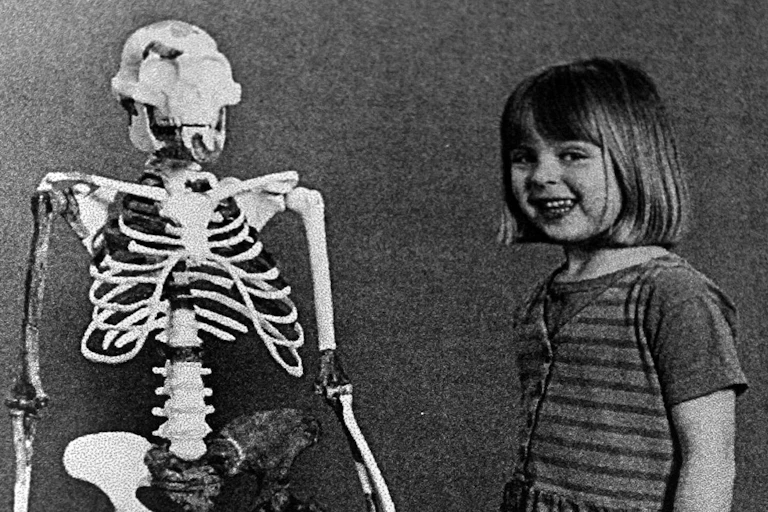 Uma criança sorridente, com as mãos nos bolsos do vestido, está ao lado do esqueleto reconstruído de um humanoide primitivo. Eles têm aproximadamente a mesma altura e tamanhos de cabeça comparáveis.