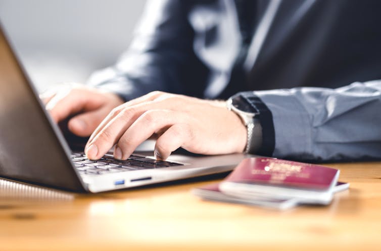 A person's hands close-up typing on a laptop with a passport next to the computer.