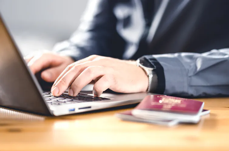 A person's hands close-up typing on a laptop with a passport next to the computer.