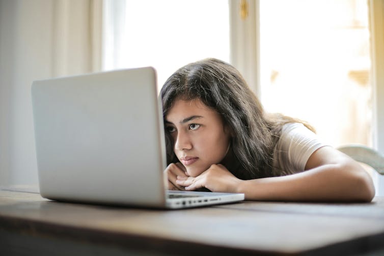 A teenage girl leans her head on a table and looks at a laptop screen.