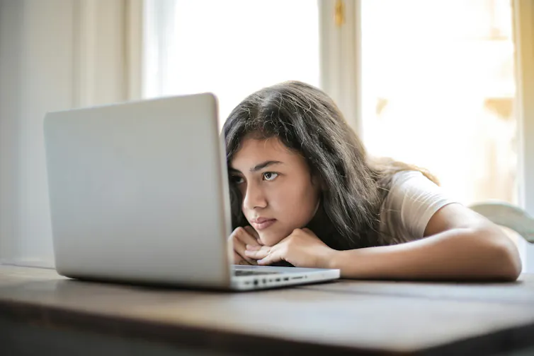 A teenage girl leans her head on a table and looks at a laptop screen.
