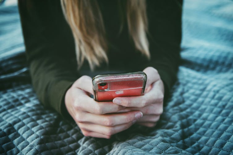 A young person lies on a bed and holds a mobile phone.