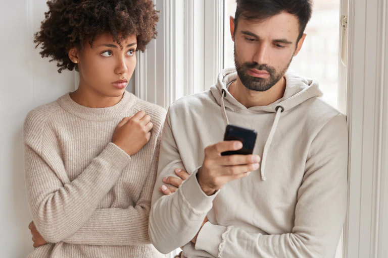Man scrolling phone with partner beside him looking sad