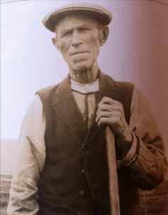 An old man dressed in farm worker garb in a sepia photograph.