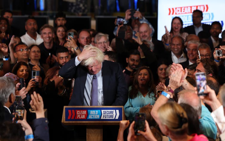 Boris Johnson scratching his head at a podium in the middle of a crowd of people.