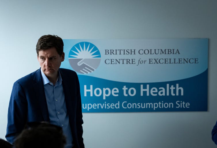 A man in a blue jacket stands next to a sign that reads, “Hope to Health Supervised Consumption Facility”