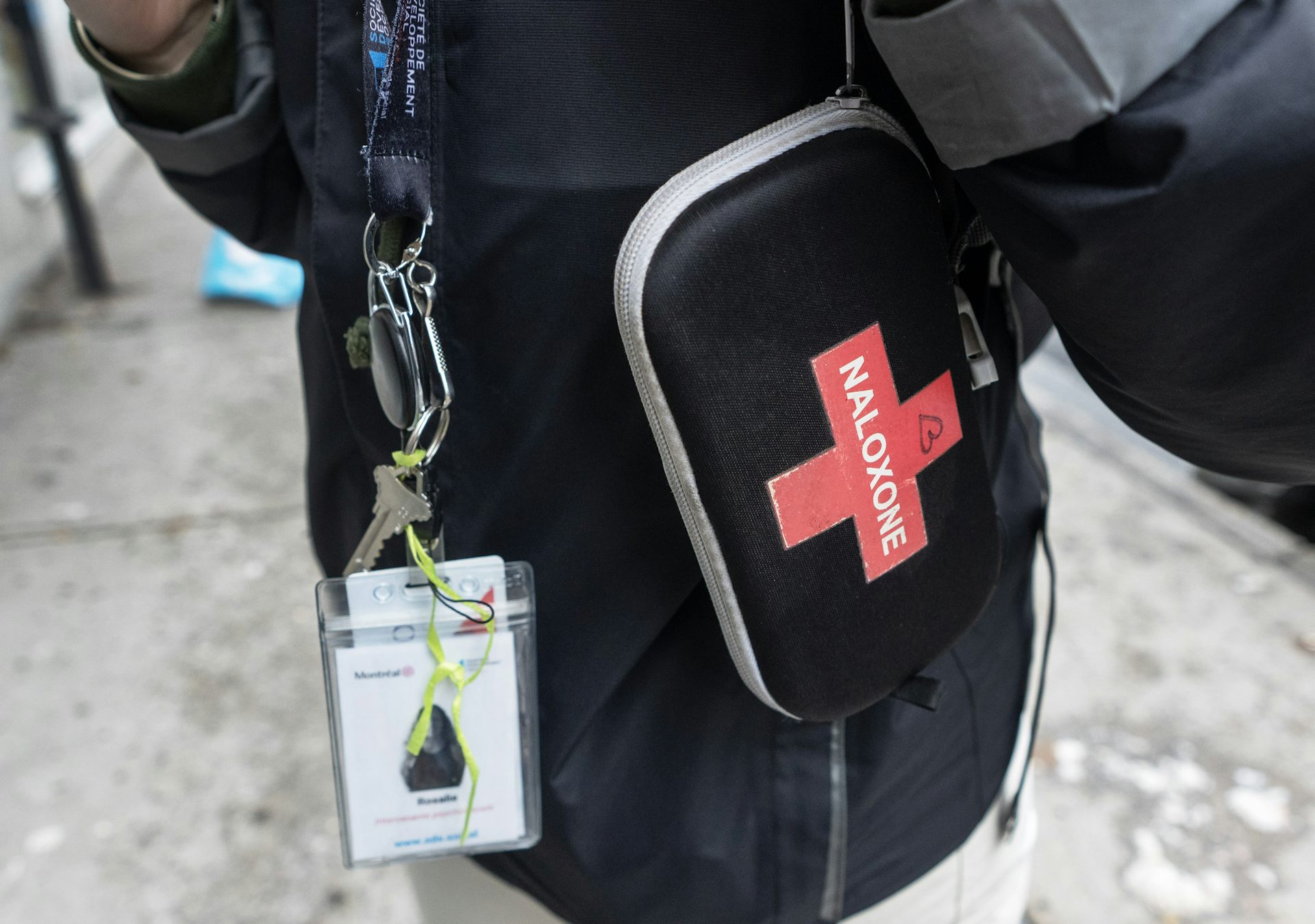Close-up photo on a black naloxone kit with a red cross on it, and a blurred ID badge on a lanyard.