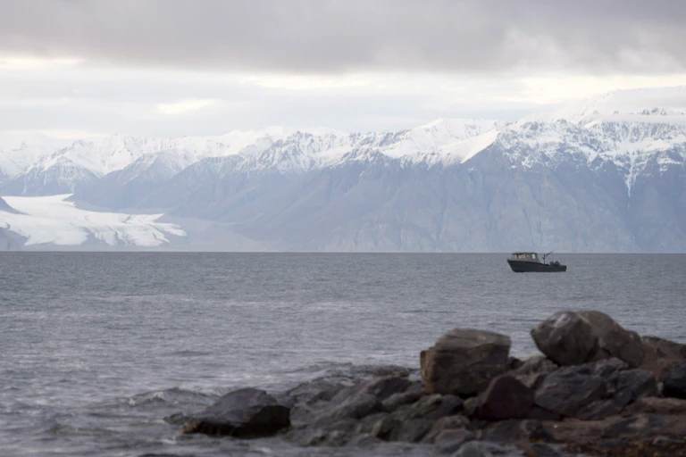 A small boat sit on the surface of a body of water, snow-capped mountains behind it.