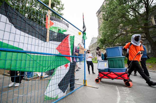 someone in an orange keffiyeh whose face is covered pulls a wagon of boxes past a Palestinian flag