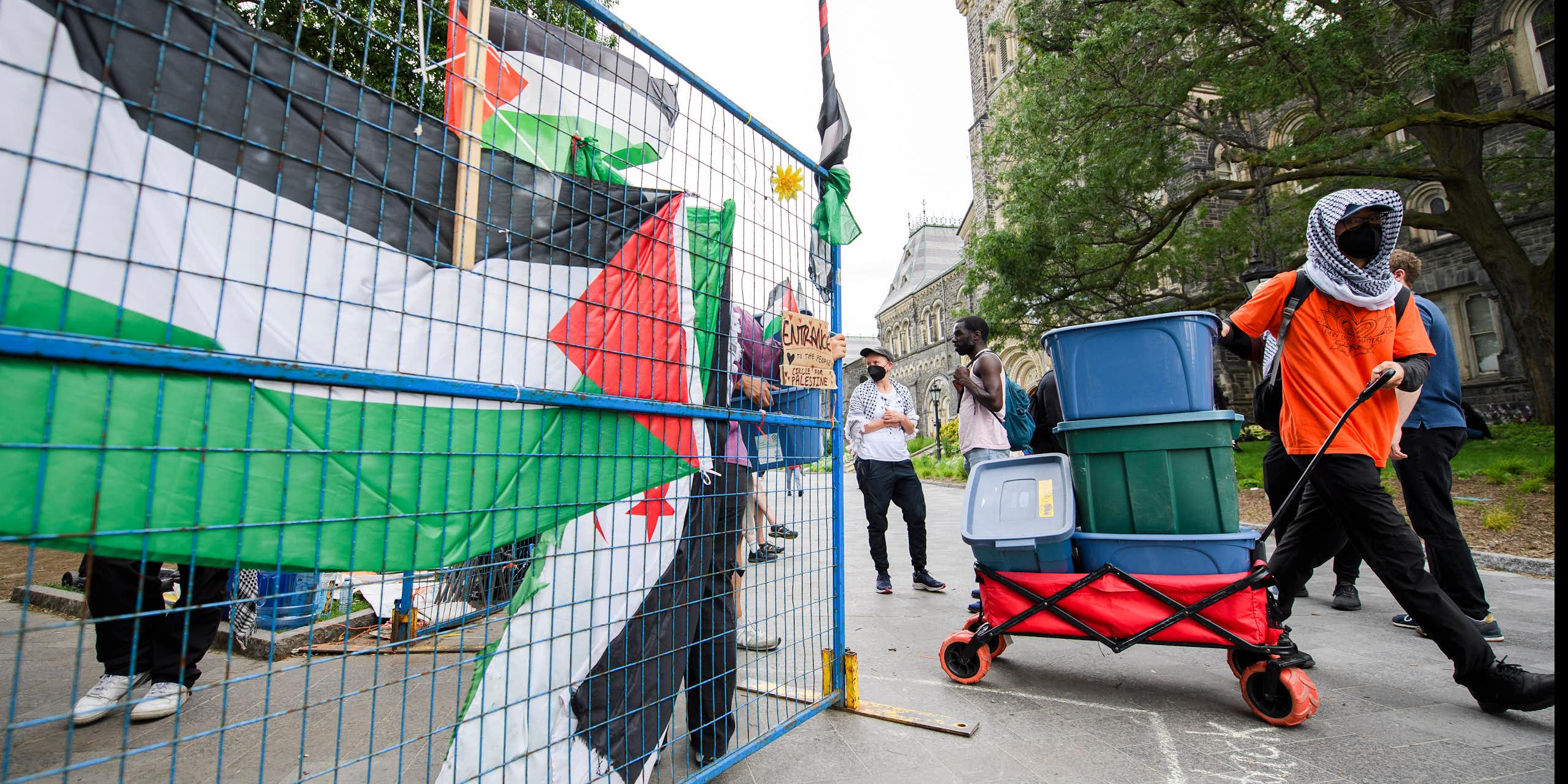 someone in an orange keffiyeh whose face is covered pulls a wagon of boxes past a Palestinian flag