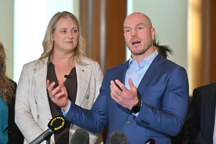 A woman stands next to a man, who's speaking at a press conference.